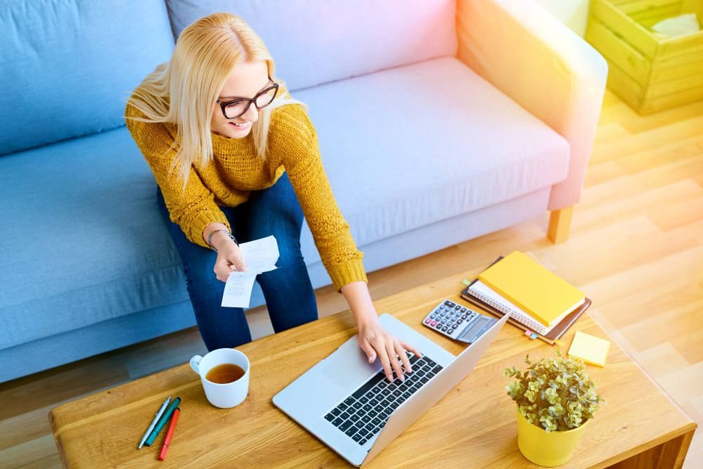 Happy young woman paying bills on laptop sitting on sofa | How To Stick To A Budget: Ways To Keep Track Of Expenses | personal expense tracker