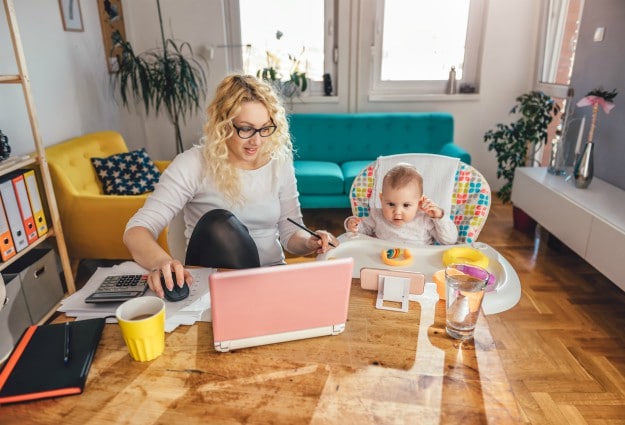 Mother wearing eyeglasses working at home office on laptop and taking care of her baby | Reasons Why Virtual Bookkeeping Is A Great Job For Stay At Home Moms | virtual bookkeeping jobs from home