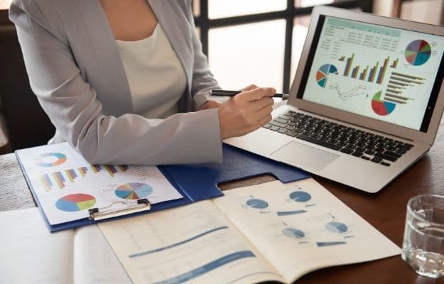 a woman is pointing at the laptop with financial data and many documents on the table ca